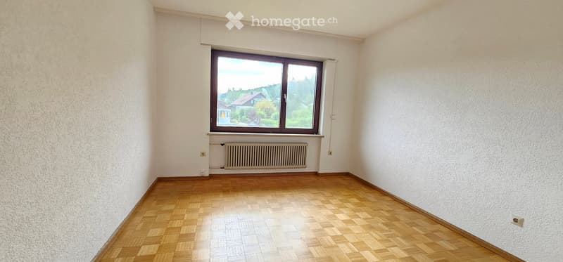 Empty room with wooden flooring, white walls, large window with view, radiator below window.