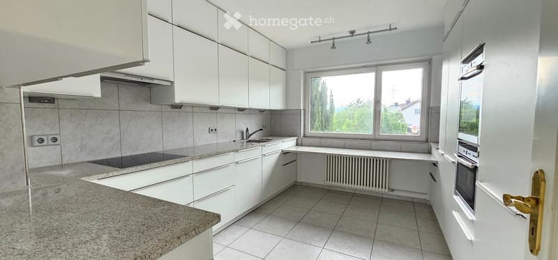 Modern kitchen with white cabinets, granite countertop, sink, oven, and radiator under a large window.