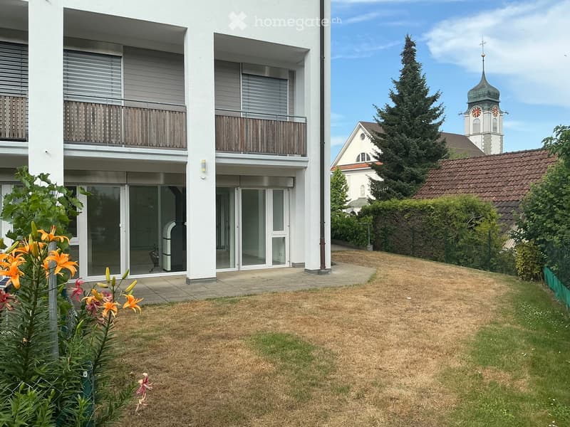 Modern two-story apartment building with balconies, glass doors, and a small garden area with flowers and grass.