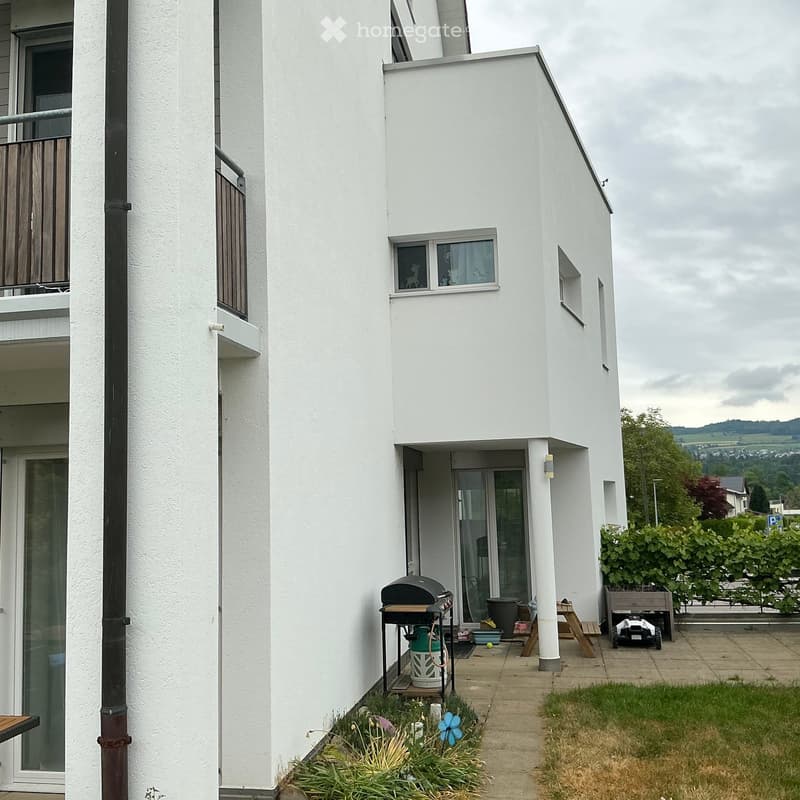 Balcony area of a modern white building with a barbecue, small garden, and scenic view of the countryside.