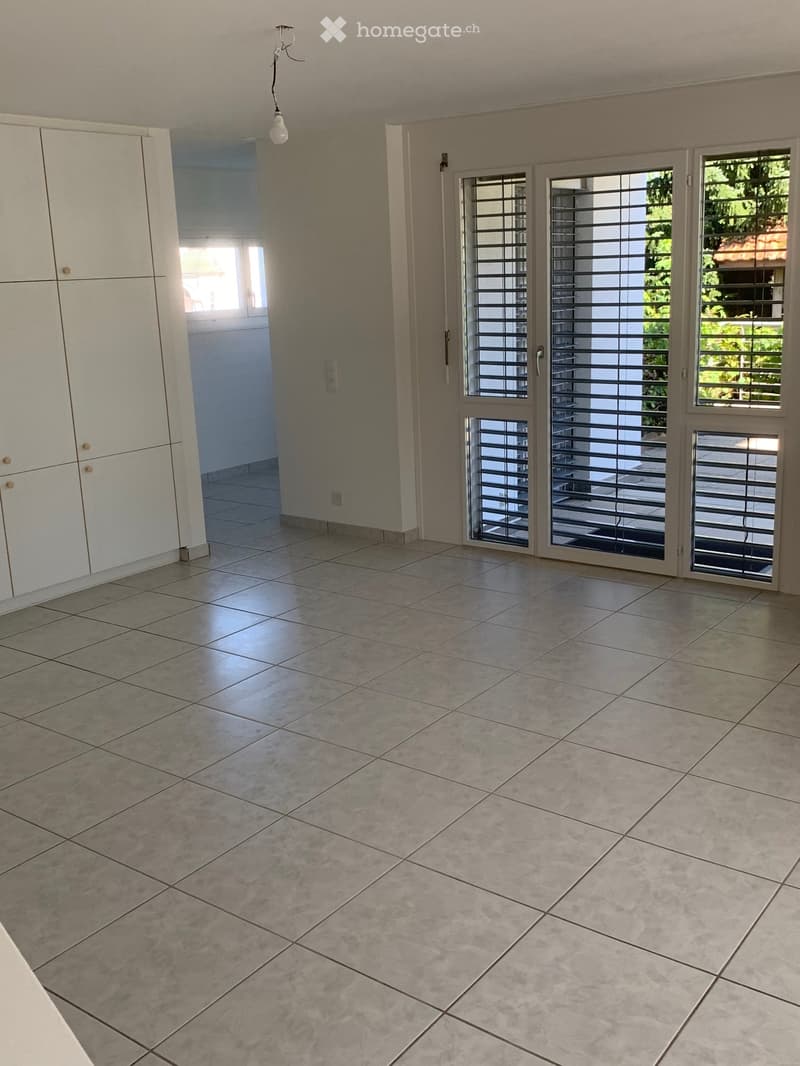 An empty room with white tiled flooring, white walls, and a white cabinet. There is a light fixture hanging from the ceiling and a glass door leading to another room. The room has a bright and clean appearance.
