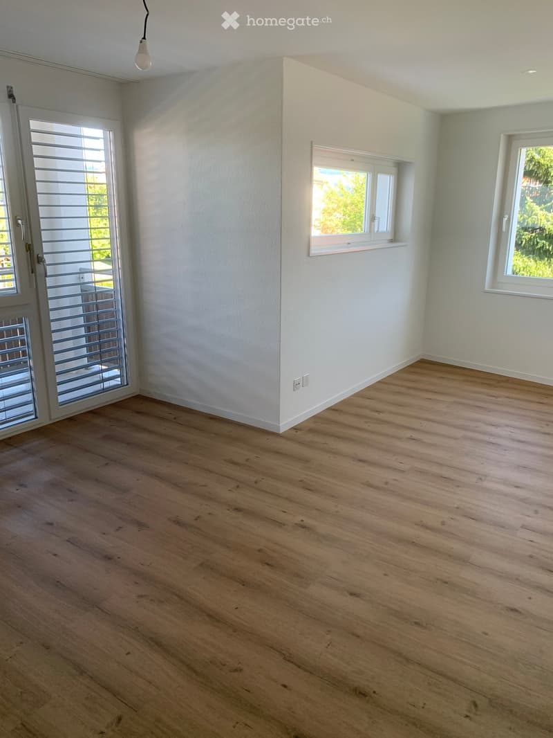 Empty room with light wooden flooring, white walls, and large windows allowing natural light. Features a sliding glass door leading to an outdoor area.