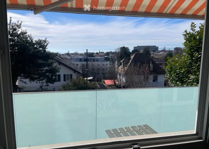 Balcony with glass railing, offering a view of a cityscape with buildings, trees, and a distant body of water under a partly cloudy sky.
