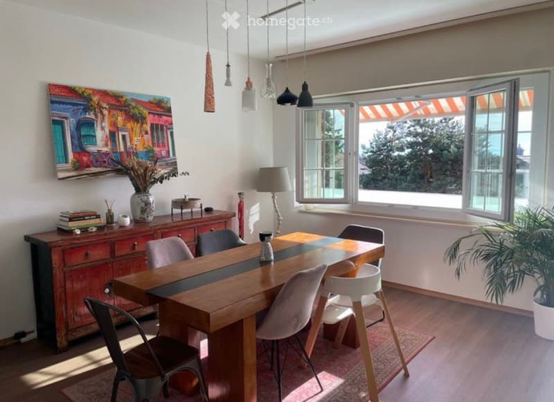Bright dining area with a long wooden table surrounded by modern chairs, a sideboard with decorative items, a lamp, and a large painting on the wall. Large windows allow natural light to fill the space.