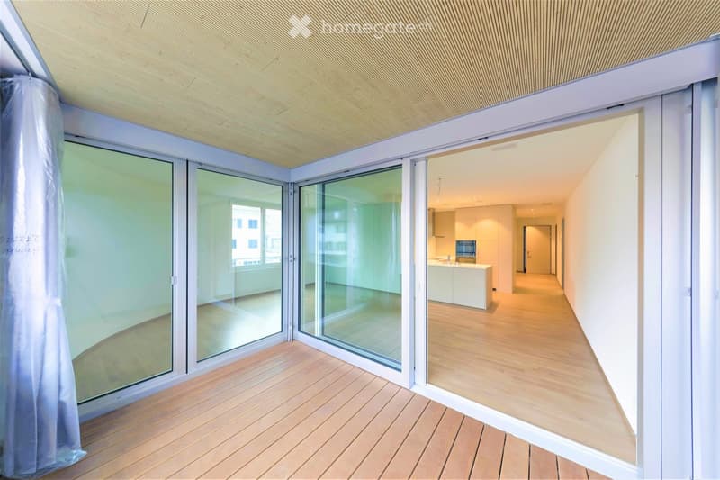 Bright, open-plan space with large glass sliding doors leading to a hallway and kitchen area. Light wooden flooring, white walls, and modern ceiling design.
