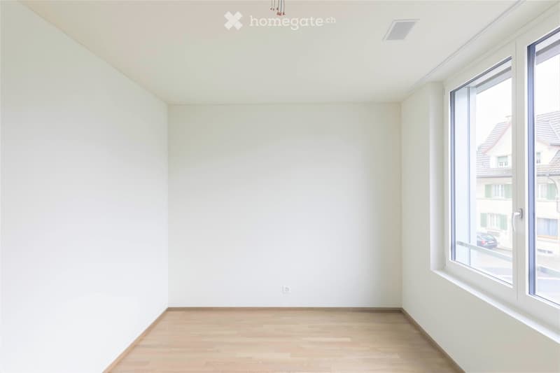 Empty room with light-colored walls and wooden flooring, featuring a large window with a view of the street.