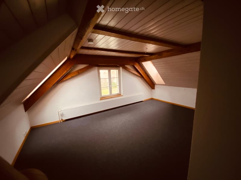 Empty attic room with wooden beams, slanted ceilings, and a single window. The floor is carpeted, and the walls are painted white.