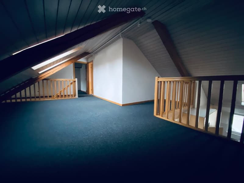 Empty attic space with wooden beams, blue carpet, and wooden railings leading to another room.