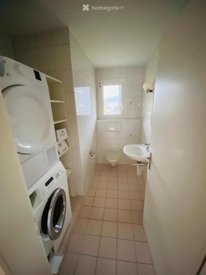 Bathroom with a toilet, sink, washing machine, dryer, and shelves. The room has tiled walls and floor, and a small window providing natural light.