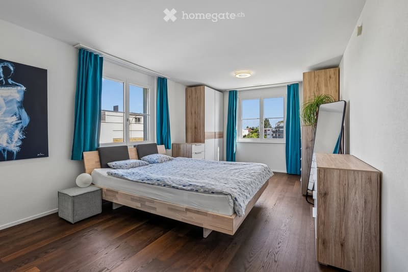 Modern bedroom with wooden flooring, large windows with blue curtains, a wooden bed with pillows and a patterned duvet, a nightstand with a decorative sphere, a wardrobe, and a potted plant near the window.