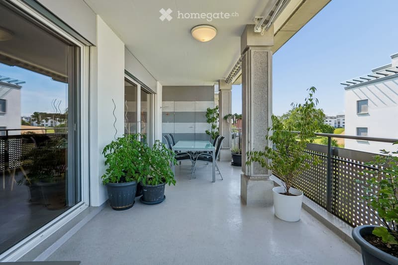Modern balcony with glass doors, concrete floor, and potted plants, featuring a small table and chairs.