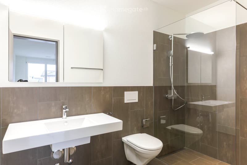 Modern bathroom with a white sink, toilet, glass-enclosed shower, and a large mirror above the sink. The walls are tiled in brown and beige, and there is a window providing natural light.
