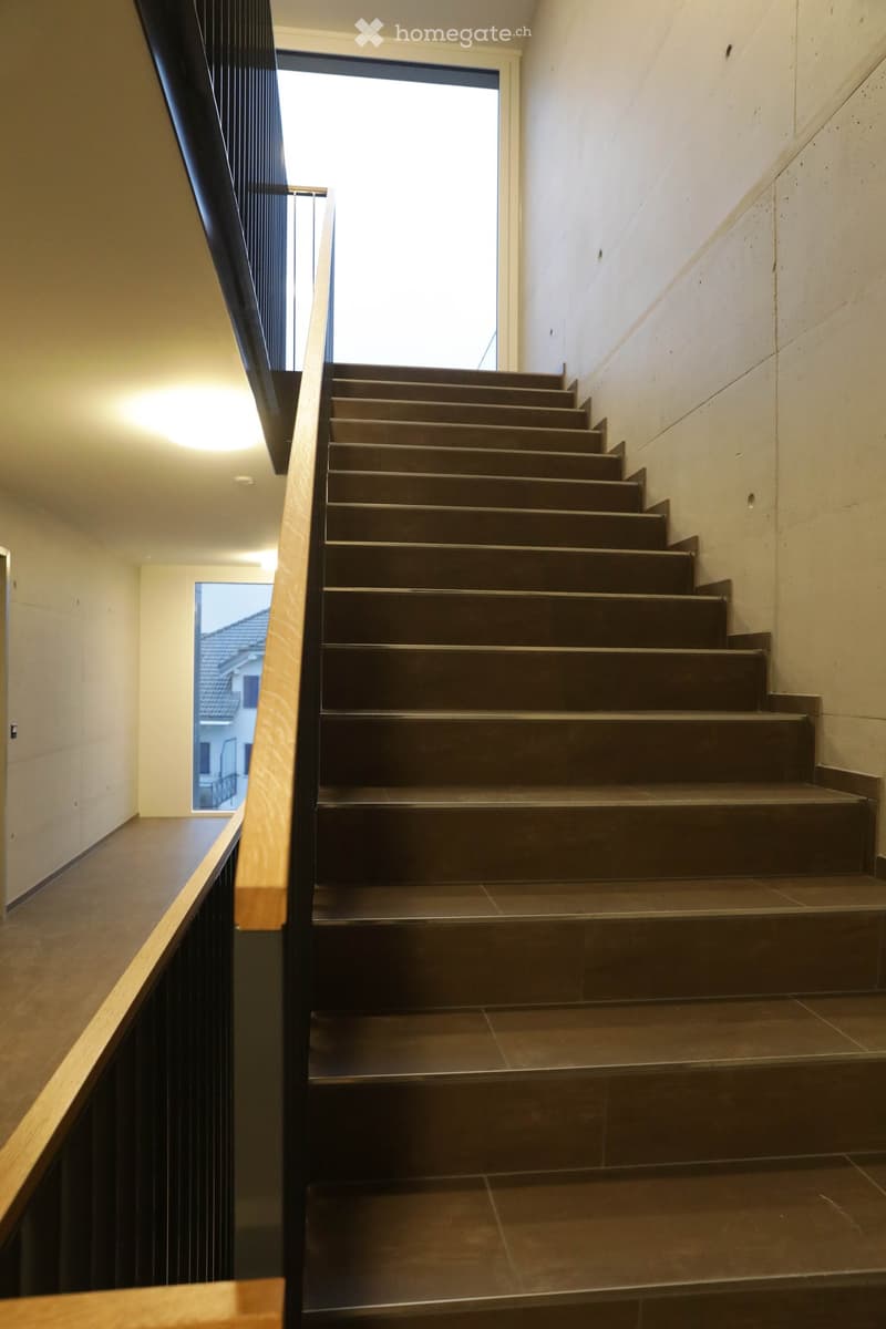 Modern staircase with dark brown steps and a wooden handrail, illuminated by recessed lighting, with a view of a neighboring building through a window.