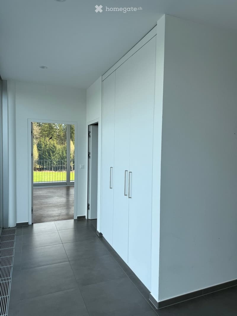 Modern hallway with white walls, dark tiled floor, built-in white storage units, and an open doorway leading to a balcony area with outdoor decking and greenery visible.