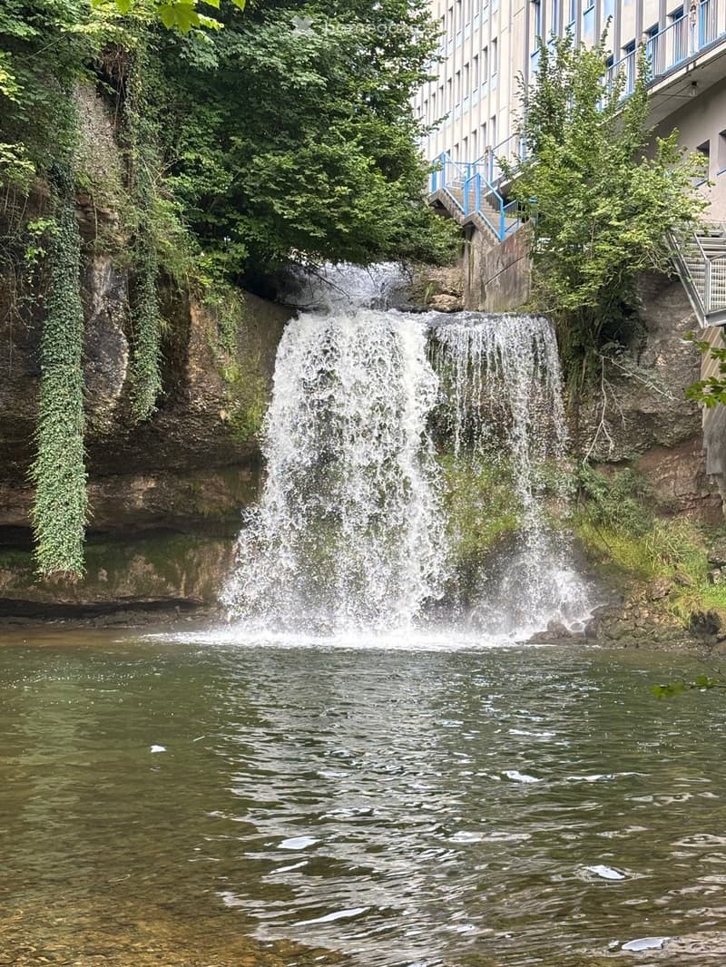 A small waterfall cascades over a rocky ledge into a calm pool below, surrounded by lush greenery and a modern building with a staircase in the background.