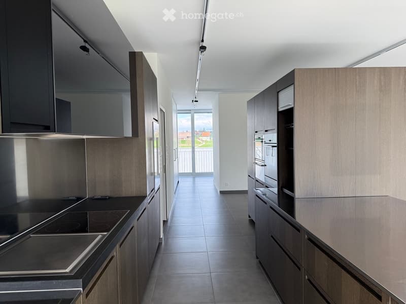 Modern kitchen with wooden cabinets, black induction cooktop, and stainless steel refrigerator. Large window offers a view of the outdoor area.