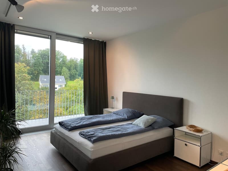 Modern bedroom with a large bed, nightstand, lamp, and a view of a green landscape through large windows.