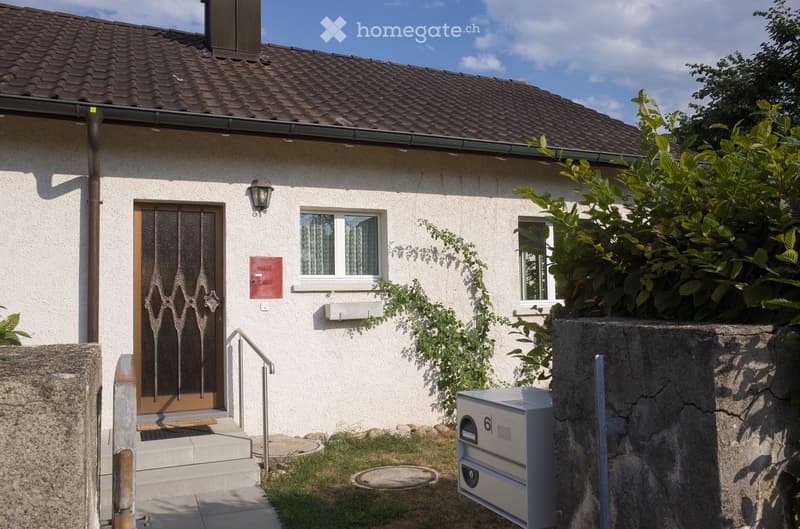 Single-story house with white exterior, brown tiled roof, and decorative front door with glass panels. Features include a small porch, a window with white frame, and a mailbox.
