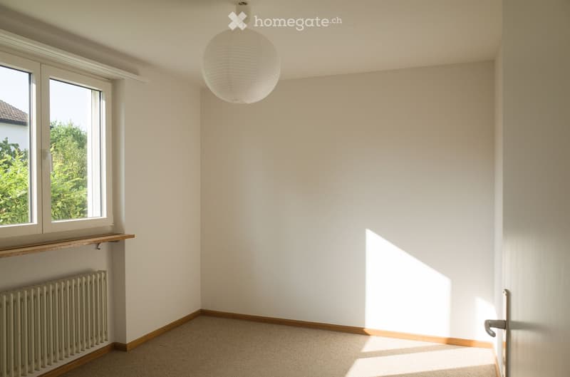 Empty room with white walls, wooden baseboards, carpeted floor, large window with wooden shelf, radiator, and a hanging spherical light fixture.