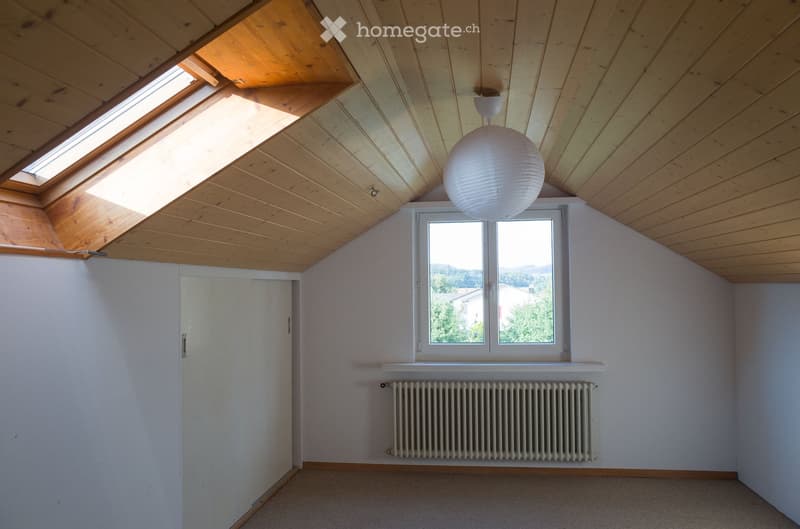 Empty attic room with wooden slanted ceilings, skylights, large window, radiator, and a hanging paper lantern light fixture.