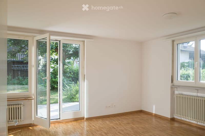 Empty room with wooden flooring, white walls, large windows, and radiators. The room has a sliding glass door leading to a garden area.