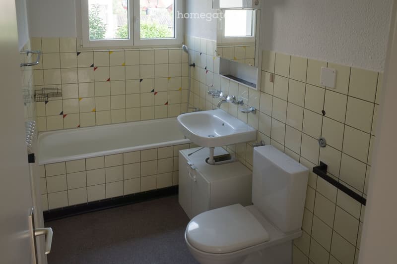 Bathroom with a bathtub, sink, toilet, mirror, and cabinet. The walls are tiled with a mix of light and dark tiles, and there are two windows providing natural light.