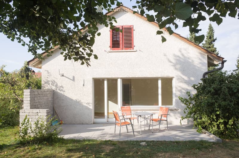A modern single-story house with white walls and red shutters, featuring a small patio area with a table and chairs, surrounded by greenery and a stone wall.