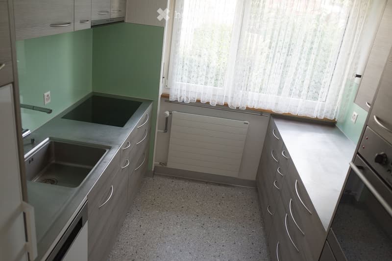 Modern kitchen with grey cabinets, stainless steel sink, and built-in oven. Green backsplash and white curtains on the window.