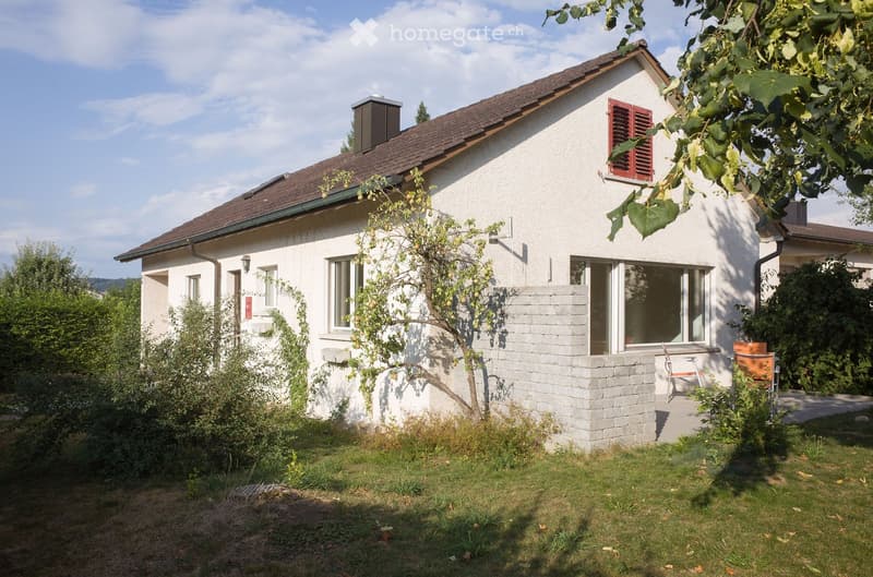 Single-story white house with a brown tiled roof, multiple windows, and a small chimney. The house is surrounded by a well-maintained garden with grass, shrubs, and a small tree. A brick patio area with outdoor furniture is visible near the entrance.