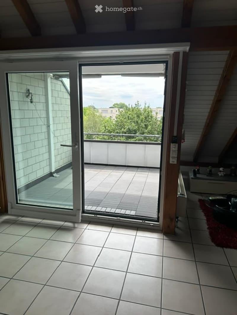 Bright, unfurnished room with large sliding glass doors opening to a balcony. The room features white tiled flooring and white walls, with exposed wooden beams on the ceiling.