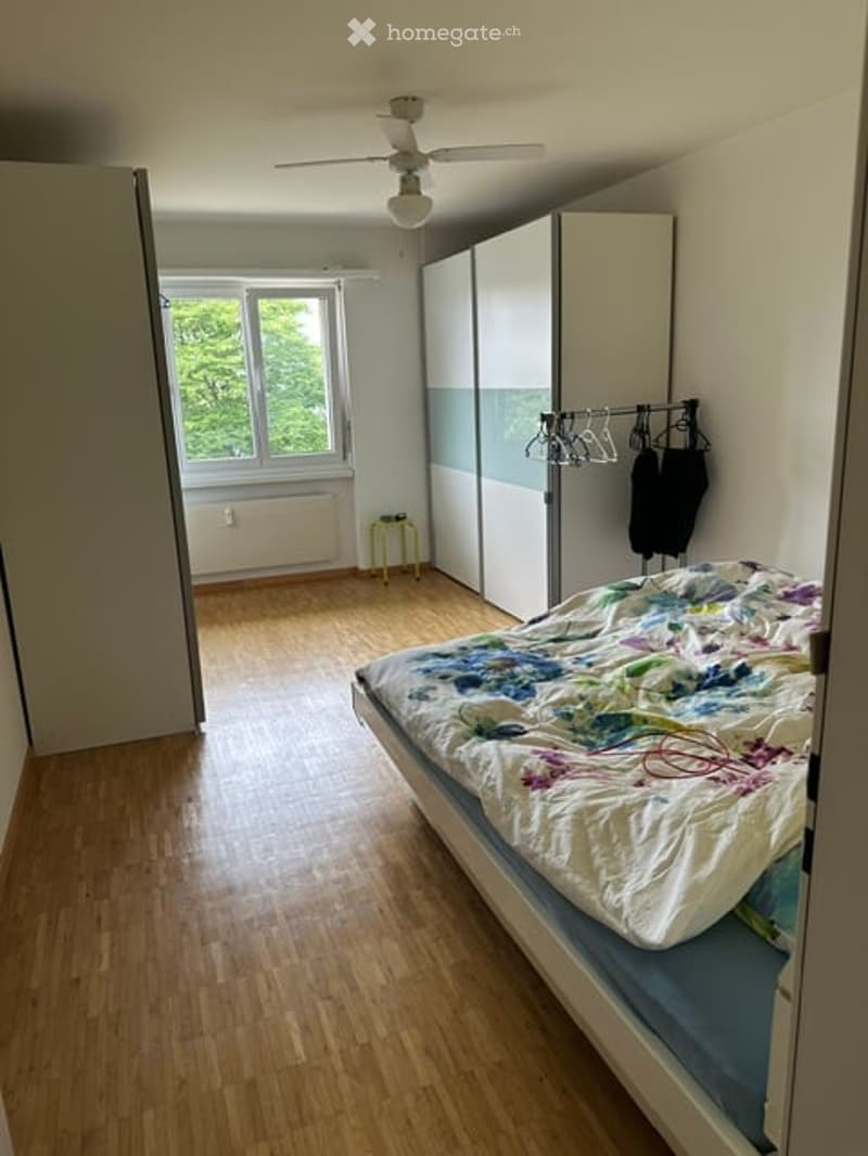 Bedroom with wooden flooring, a bed with a colorful duvet, a wardrobe, and a small stool near the window.
