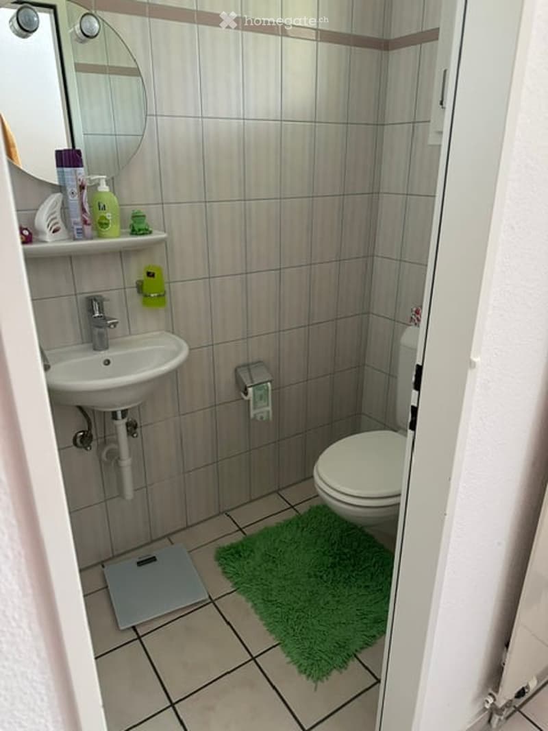 Compact bathroom with tiled walls and floor, a white sink with a mirror above it, a toilet, a green bath mat, and a scale on the floor. Shelves hold shampoo, conditioner, and body wash bottles.