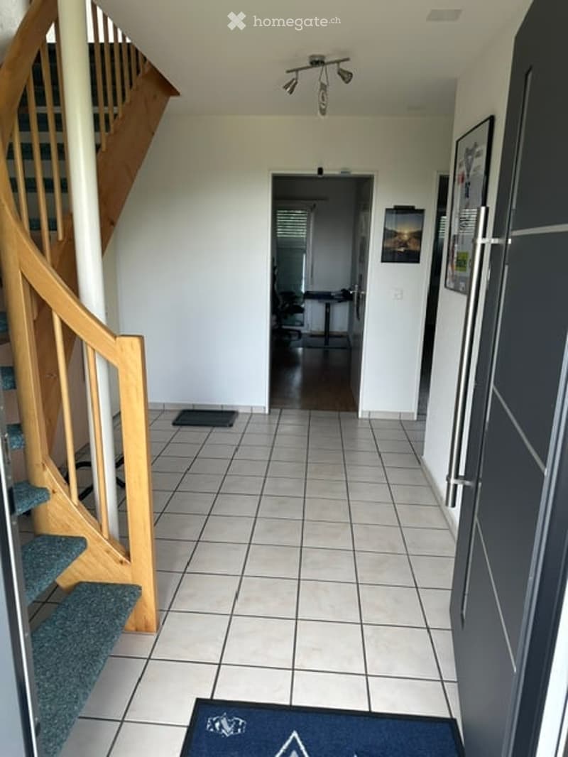 Clean, modern hallway with tiled flooring, wooden staircase, and a view into an adjacent room with a table and chairs.
