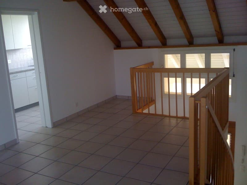 Empty room with tiled floor, wooden staircase railing, and exposed wooden beams on the ceiling. Natural light from windows and an open doorway to another room.