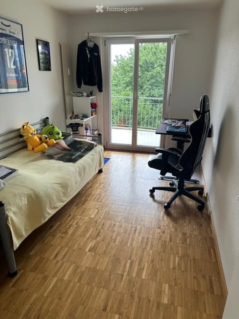 Cozy bedroom with wooden flooring, a bed with a yellow blanket, a laptop on the bed, a gaming chair, a small table, and a balcony with a view of greenery.