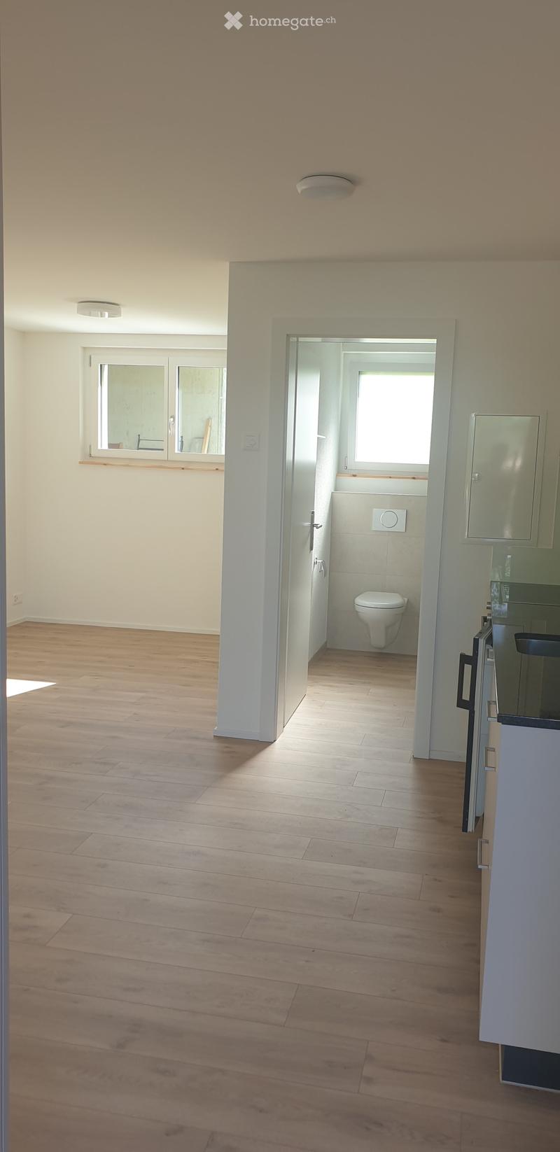 Empty room with wooden flooring, white walls, and a doorway leading to a small bathroom with a toilet and window.