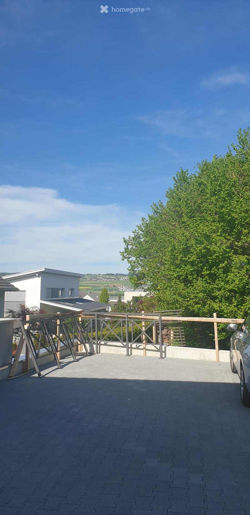Balcony with wooden railing, paved floor, and a view of a house and greenery in the background.