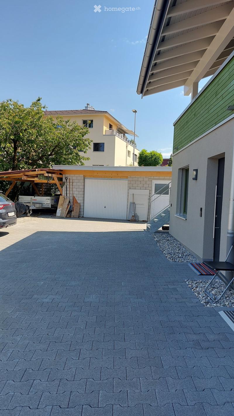Outdoor patio area with paved stone tiles, a covered wooden structure, a garage, and a modern house in the background.