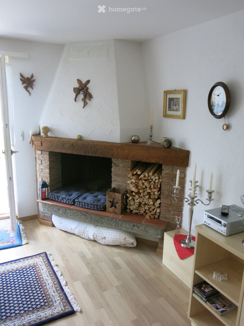 Living room with a brick fireplace, wooden mantel, decorative items, and a stack of firewood. Includes a shelf with books and a clock on the wall.