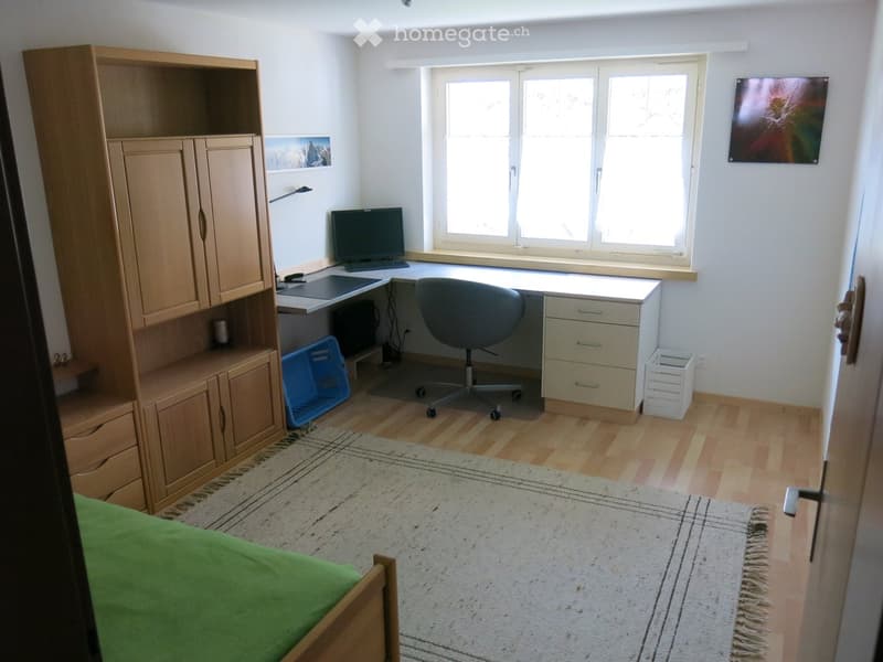Well-lit room with a desk setup, a bed, and a large window letting in natural light. The room features a wooden cabinet, a computer, a chair, and a colorful artwork on the wall.