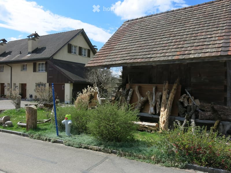 Two-story house with a tiled roof, beige walls, and multiple windows. A wooden outbuilding with a slanted roof is visible, along with a stack of firewood and some greenery in the front yard.
