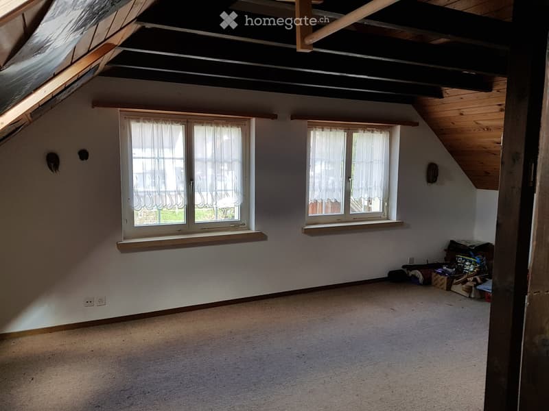 Empty attic room with wooden beams, skylight, and two windows with white curtains. Bare walls and carpeted floor.