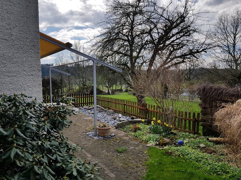 Garden area with a paved pathway, wooden fence, potted plants, and a covered patio area attached to a building.