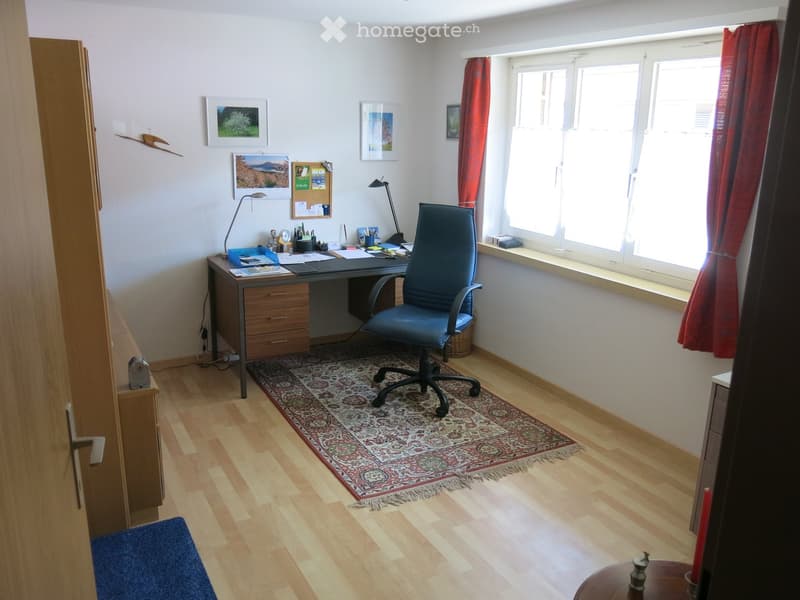 A well-lit office space with a wooden floor, a large window with red curtains, a desk with a chair, a lamp, and a patterned rug.