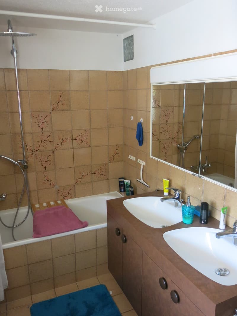 Bathroom with a bathtub, double sink, mirror, and shower area. Brown tiled walls and wooden vanity.