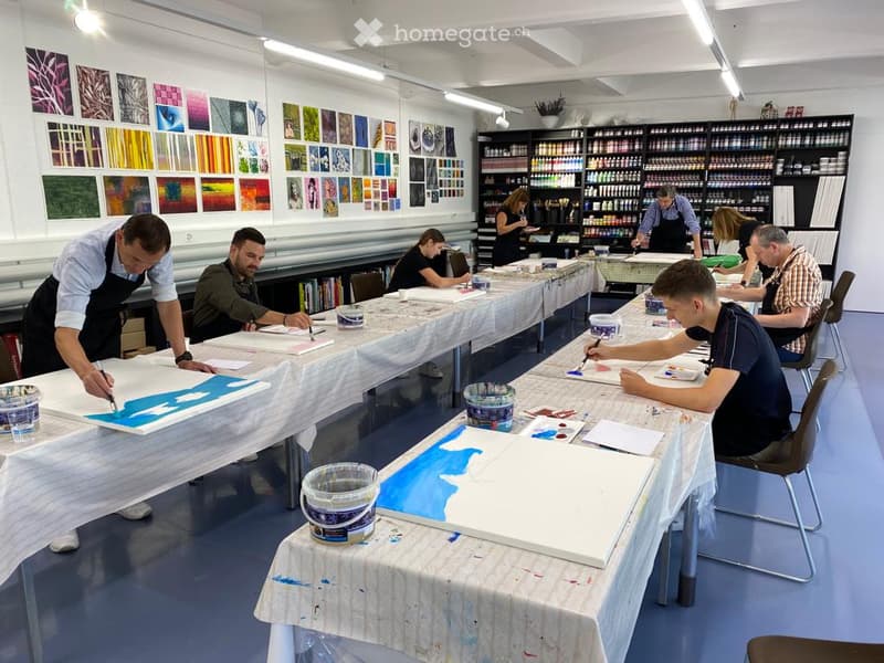 Art studio with multiple tables covered in white cloths, people painting on canvases, shelves filled with art supplies, and colorful paintings displayed on the walls.