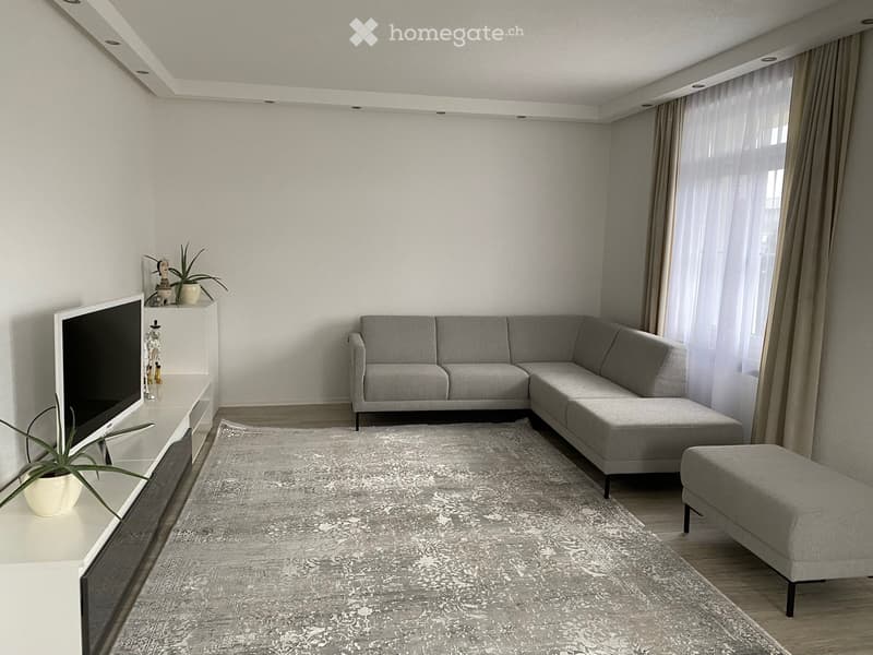 Modern living room with grey sectional sofa, ottoman, flat-screen TV on a white stand, and decorative plants.