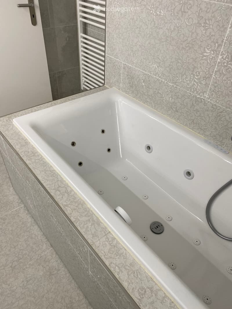 Modern bathroom with a white bathtub featuring multiple jets, surrounded by grey marble tiles and a white radiator.