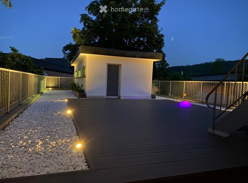 Modern outdoor deck with wooden flooring, integrated lighting, and a small white building with a grey door and windows.