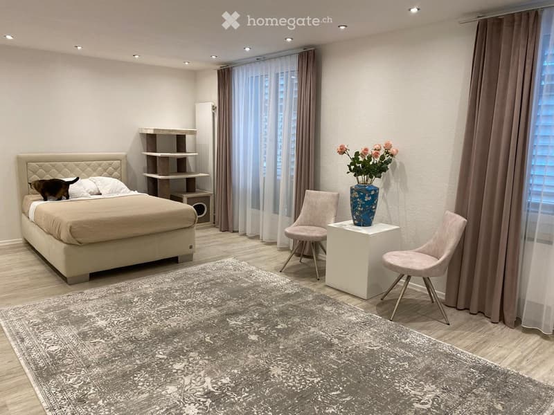 Modern bedroom with a large bed, two chairs, a small table with a vase, and a shelf unit. The room features wooden flooring, large windows with curtains, and a patterned rug.